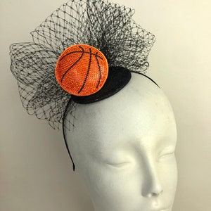 May include: A black headband with a decorative basketball-themed fascinator. The fascinator features a round, orange basketball design with black lines and a black net veil. The basketball is embellished with sparkling rhinestones.