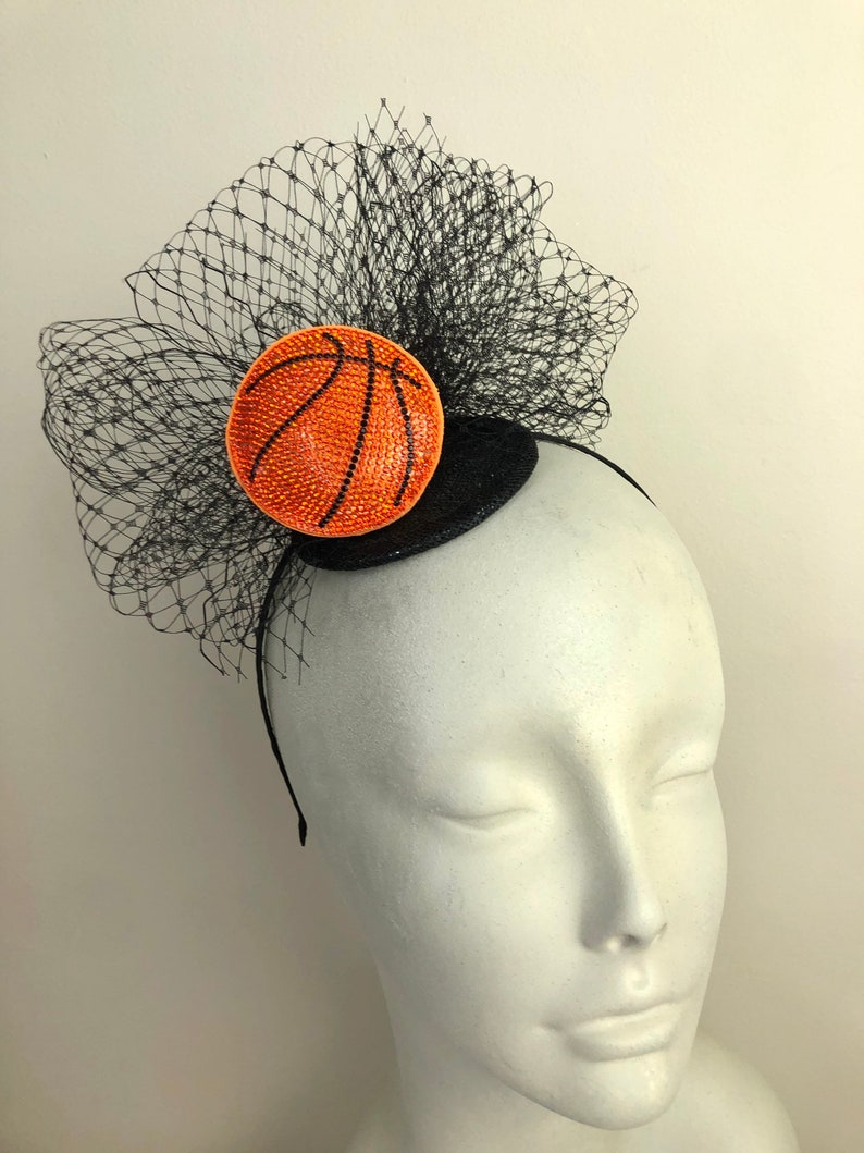 May include: A black headband with a decorative basketball-themed fascinator. The fascinator features a round, orange basketball design with black lines and a black net veil. The basketball is embellished with sparkling rhinestones.