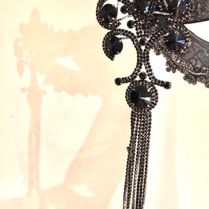 May include: A black masquerade mask with a floral design and a long chain of black beads. The mask is decorated with rhinestones.