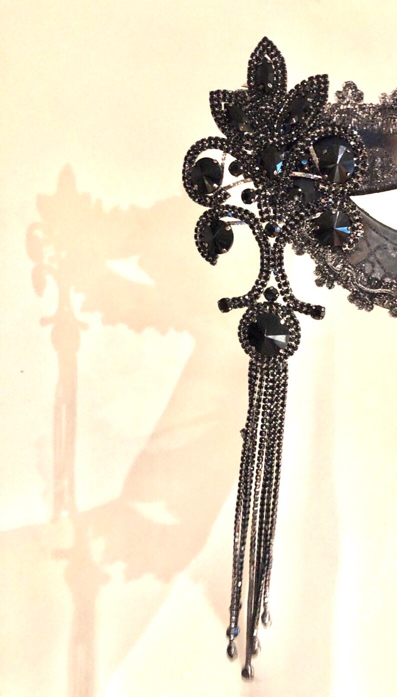May include: A black masquerade mask with a floral design and a long chain of black beads. The mask is decorated with rhinestones.