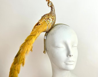 May include: A decorative headpiece featuring a golden peacock with a glittered body and long, flowing feathers. The peacock sits atop a thin, gold headband. The headpiece is displayed on a white mannequin head.