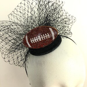 May include: A black headpiece with a football-shaped embellishment. The football is brown with white accents and covered in rhinestones. A black net veil and a black base complete the design.