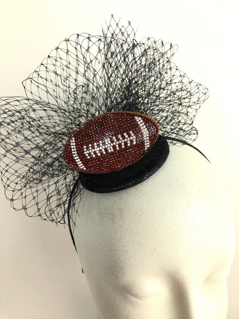 May include: A black headpiece with a football-shaped embellishment. The football is brown with white accents and covered in rhinestones. A black net veil and a black base complete the design.