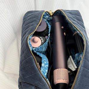 May include: A dark blue corduroy zippered pouch with a gold-toned zipper, lined with a blue and white floral fabric. The pouch contains a dark purple and pink hair styling tool with multiple attachments and a black power cord inside the main compartment.