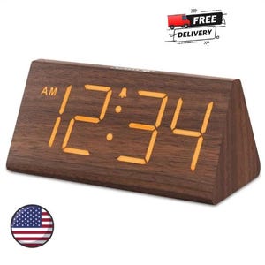 May include: A triangular digital alarm clock with a dark brown wood grain finish. The front face displays the time 12:34 in bright orange LED numerals, including an AM indicator and a small bell icon for the alarm setting on the display.
