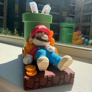 May include: A 3D-printed desk organizer featuring a sleeping Mario figure resting against a green pipe. The base is brown with a brick-like texture, and two yellow coins lie on the surface. Two clear pens with red ink are stored inside the green pipe.