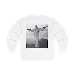 May include: A white long-sleeve shirt featuring a black and white graphic print on the chest. The graphic depicts the Christ the Redeemer statue with arms outstretched, holding a submachine gun in each hand against a mountain landscape background.