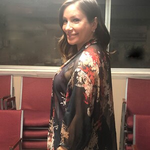 Puede incluir: Un kimono floral negro transparente con detalles en rojo y blanco. La tela ligera cae sobre los hombros y los brazos. El kimono se lleva sobre una parte superior oscura y se combina con pantalones negros.