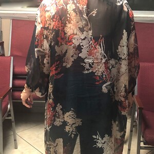 Puede incluir: Un kimono negro largo y transparente con un estampado floral en tonos rojos, blancos y beige. El kimono se lleva sobre pantalones oscuros y una camiseta. Las mangas son largas y fluidas, y la tela parece ligera.