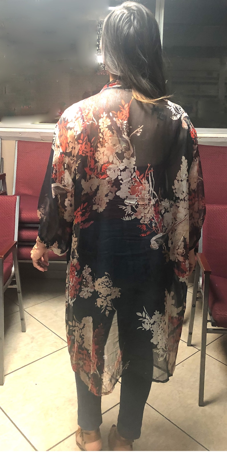Puede incluir: Un kimono negro largo y transparente con un estampado floral en tonos rojos, blancos y beige. El kimono se lleva sobre pantalones oscuros y una camiseta. Las mangas son largas y fluidas, y la tela parece ligera.