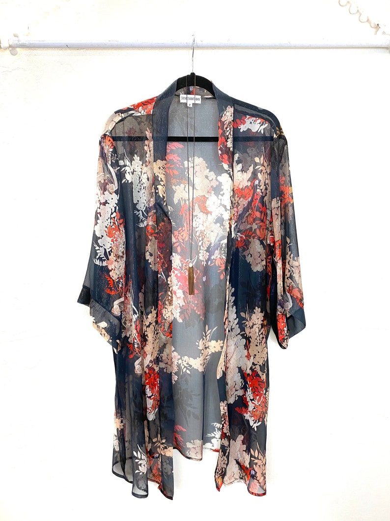 Puede incluir: Un kimono transparente con frente abierto y un estampado floral en tonos azul marino, rojo y crema. El kimono est&aacute; colgado de una percha negra sobre un fondo blanco. Se ve un collar largo.