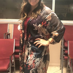 Puede incluir: Una parte superior tipo kimono oscura y transparente con un estampado floral en rojo, blanco y amarillo. Se usa sobre una parte superior roja y vaqueros azules. El fondo incluye sillas rojas y una ventana.