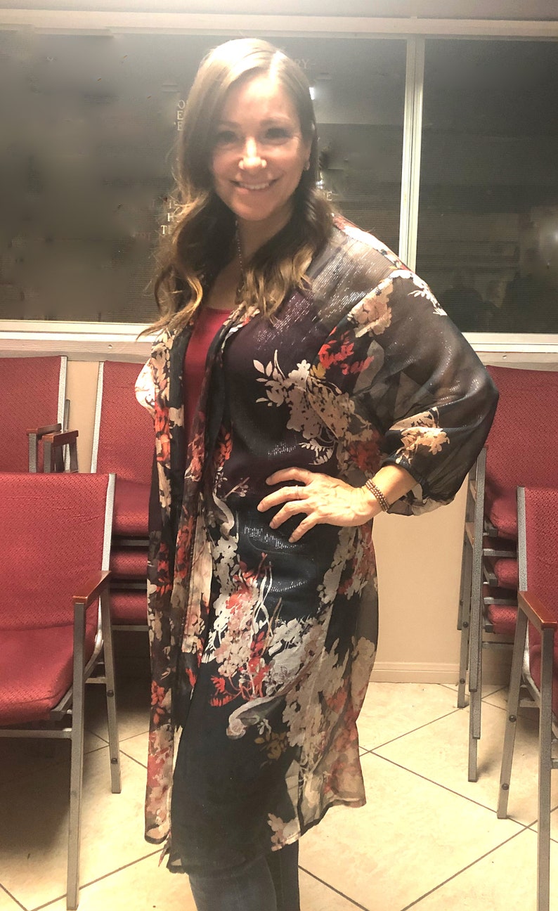 Puede incluir: Una parte superior tipo kimono oscura y transparente con un estampado floral en rojo, blanco y amarillo. Se usa sobre una parte superior roja y vaqueros azules. El fondo incluye sillas rojas y una ventana.