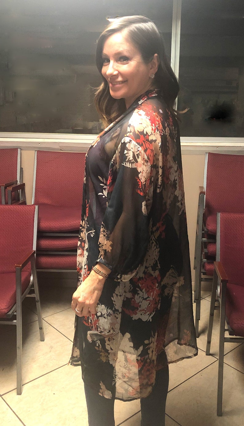 Puede incluir: Un kimono floral negro transparente con detalles en rojo y blanco. La tela ligera cae sobre los hombros y los brazos. El kimono se lleva sobre una parte superior oscura y se combina con pantalones negros.