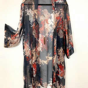 Puede incluir: Un kimono transparente de frente abierto con un estampado floral en tonos azul marino, rojo y beige. El kimono est&aacute; colgado de una percha negra, mostrando su dise&ntilde;o fluido y su delicado tejido. Un collar largo con borla cuelga en el centro.