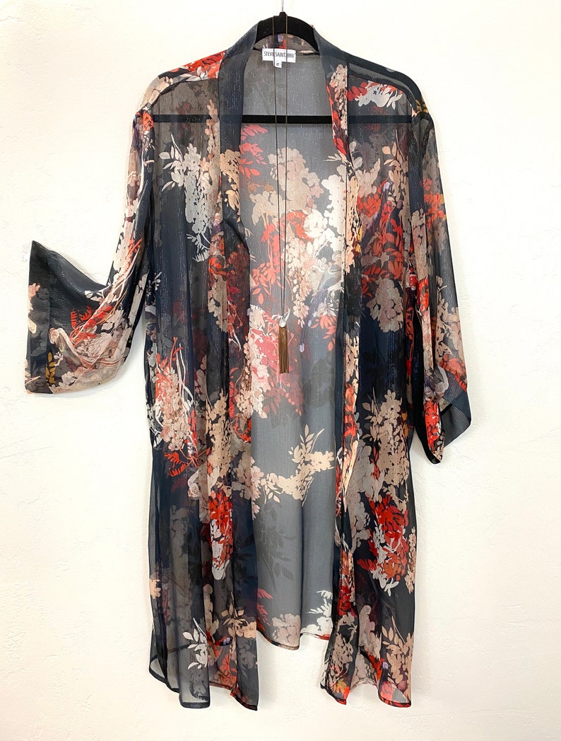 Puede incluir: Un kimono transparente de frente abierto con un estampado floral en tonos azul marino, rojo y beige. El kimono est&aacute; colgado de una percha negra, mostrando su dise&ntilde;o fluido y su delicado tejido. Un collar largo con borla cuelga en el centro.