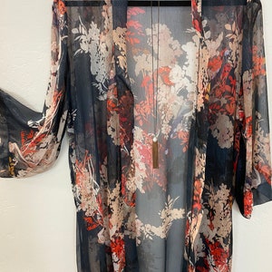 Puede incluir: Quimono transparente de frente abierto con un estampado floral en tonos rojos, crema y beige sobre un fondo azul marino oscuro. El quimono tiene mangas largas y una silueta fluida. Se ve un collar largo con borla.