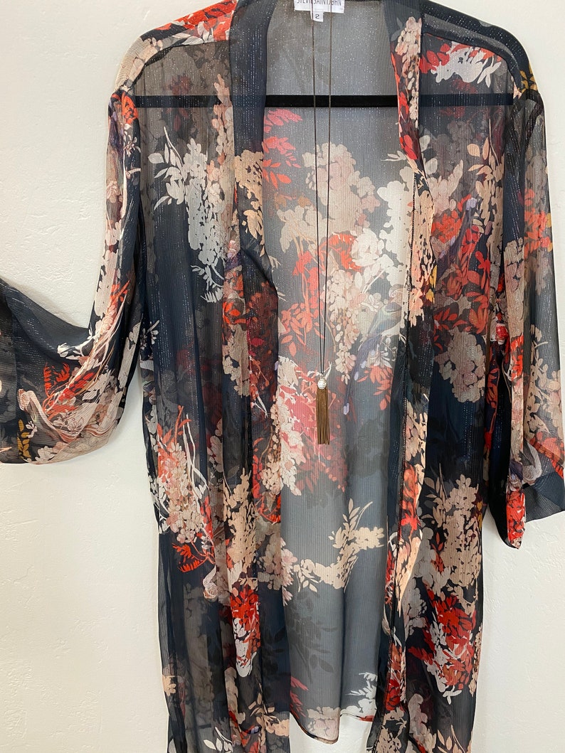 Puede incluir: Quimono transparente de frente abierto con un estampado floral en tonos rojos, crema y beige sobre un fondo azul marino oscuro. El quimono tiene mangas largas y una silueta fluida. Se ve un collar largo con borla.