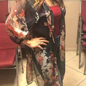 Puede incluir: Un kimono oscuro y transparente con un estampado floral en rojo, blanco y naranja. Se lleva sobre una camiseta roja y vaqueros azules. Las mangas son largas y fluidas, y el estilo general es informal y elegante.