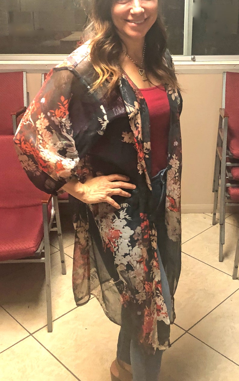 Puede incluir: Un kimono oscuro y transparente con un estampado floral en rojo, blanco y naranja. Se lleva sobre una camiseta roja y vaqueros azules. Las mangas son largas y fluidas, y el estilo general es informal y elegante.