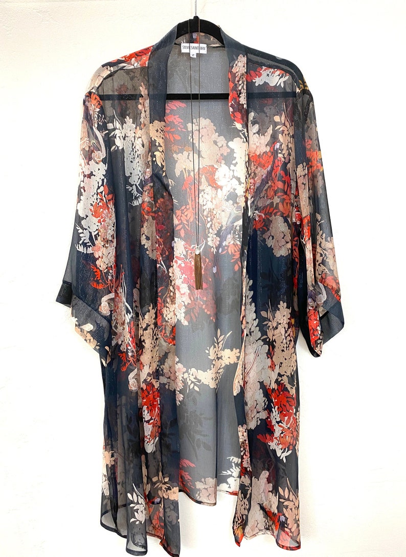 Puede incluir: Un kimono transparente con frente abierto y un estampado floral en tonos rojos, blancos y beige sobre un fondo azul marino. El kimono est&aacute; colgado de una percha negra. Tambi&eacute;n se ve un collar largo con borla.