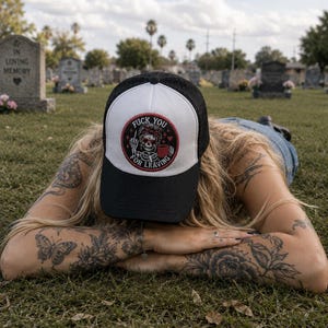 May include: A black and white trucker hat featuring a circular patch on the front. The patch displays a skeleton graphic with a red mug, surrounded by the text FUCK YOU FOR LEAVING in a circular red border with small star and heart accents.