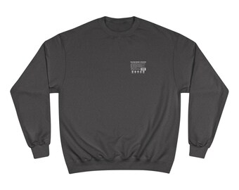 May include: A dark charcoal gray crewneck sweatshirt featuring long sleeves and ribbed cuffs. On the upper left chest, there is a small white rectangular graphic containing printed text and a barcode, resembling a nutrition facts label.