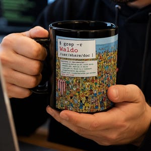 May include: A black ceramic mug featuring a graphic print of a crowded beach scene with a white text box overlay. The text reads $ grep -r Waldo /usr/share/doc followed by a list of file paths and the final line stating Waldo not found.