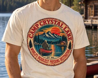 May include: A cream-colored short-sleeve t-shirt featuring a circular graphic of a person in a red canoe fishing on a lake with mountains and a cabin in the background. The text reads Camp Crystal Lake World's Deadliest Phishing Destination Est. 1935.
