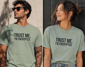 May include: A sage green short-sleeved t-shirt featuring the black text "TRUST ME, I'M ENCRYPTED." printed in a bold, sans-serif font across the chest. The garment has a classic crew neckline and a standard fit, made of a solid-colored fabric.