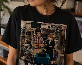 May include: A black t-shirt featuring a graphic print of two characters in an office setting with a small black box on a blue-covered table. The text "This Jen, is the internet." is printed at the bottom of the graphic, along with "I heart energy".