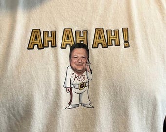 May include: A cream-colored t-shirt featuring a graphic of a person dressed in a white jumpsuit with a belt and the word King written on the chest. Above the figure, the text AH AH AH! is printed in gold letters with a black outline.