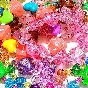 Op de afbeelding: Een kleurrijke ketting gemaakt van verschillende kralen, waaronder harten, dobbelstenen en andere vormen. De ketting is gemaakt van helder, roze en paars plastic. De dobbelstenen zijn genummerd van 1 tot 6 en van 1 tot 20. De ketting is een leuke en speelse accessoire.
