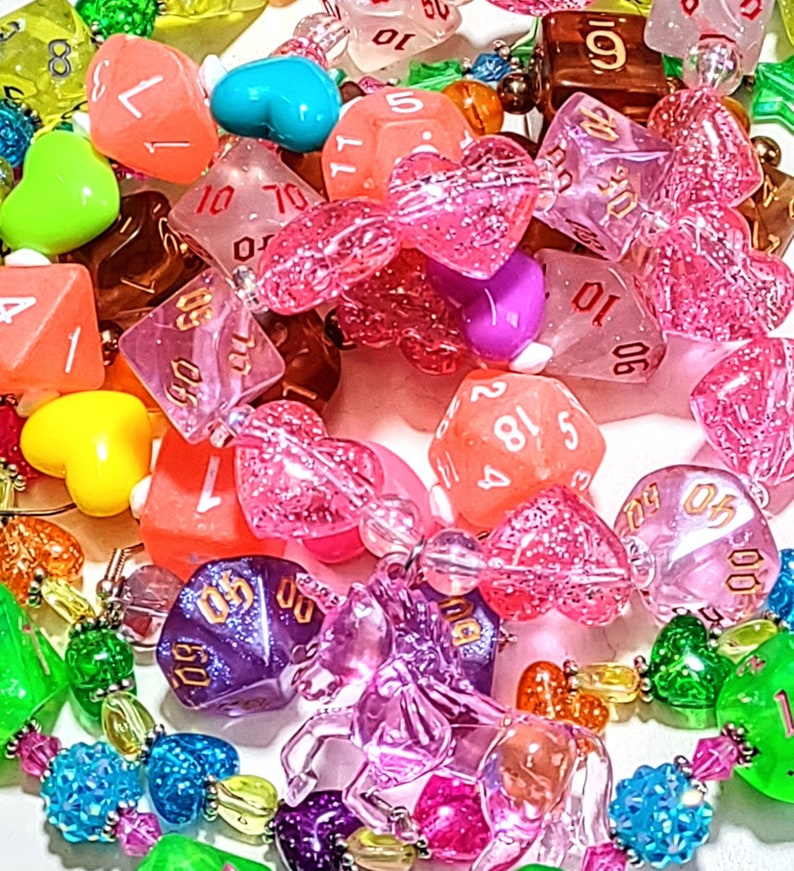 Op de afbeelding: Een kleurrijke ketting gemaakt van verschillende kralen, waaronder harten, dobbelstenen en andere vormen. De ketting is gemaakt van helder, roze en paars plastic. De dobbelstenen zijn genummerd van 1 tot 6 en van 1 tot 20. De ketting is een leuke en speelse accessoire.