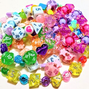Op de afbeelding: Een kleurrijke ketting gemaakt van dobbelstenen, snoepjes en andere kleine bedels. De ketting is gemaakt van verschillende kleuren, waaronder roze, blauw, groen, geel en paars. De dobbelstenen zijn allemaal van verschillende groottes en vormen, en sommige hebben nummers erop. De snoepjes zijn ook van verschillende kleuren en vormen, en sommige hebben glitter erop. De bedels zijn ook van verschillende kleuren en vormen, en sommige hebben glitter erop.