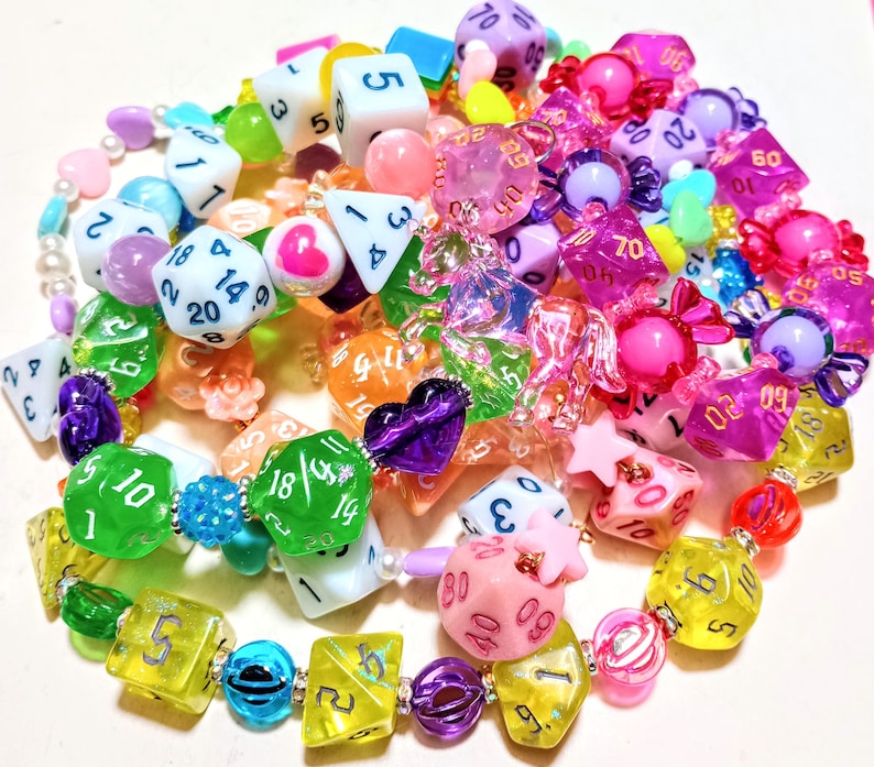 Op de afbeelding: Een kleurrijke ketting gemaakt van dobbelstenen, snoepjes en andere kleine bedels. De ketting is gemaakt van verschillende kleuren, waaronder roze, blauw, groen, geel en paars. De dobbelstenen zijn allemaal van verschillende groottes en vormen, en sommige hebben nummers erop. De snoepjes zijn ook van verschillende kleuren en vormen, en sommige hebben glitter erop. De bedels zijn ook van verschillende kleuren en vormen, en sommige hebben glitter erop.