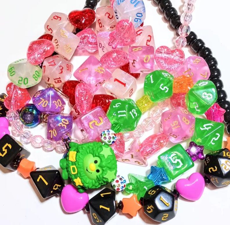 Op de afbeelding: Een kleurrijke ketting gemaakt van verschillende kralen, waaronder roze, groene en zwarte dobbelstenen, harten en sterren. De ketting heeft ook een klein groen monsterbedeltje.