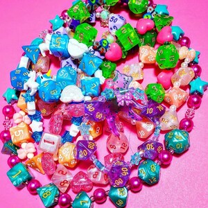Op de afbeelding: Een kleurrijke ketting gemaakt van verschillende dobbelstenen, kralen en bedels. De dobbelstenen zijn in verschillende kleuren, waaronder roze, blauw, groen en paars. De kralen zijn in verschillende kleuren, waaronder roze, wit en zwart. De bedels omvatten een vlinder, een hart en een ster.