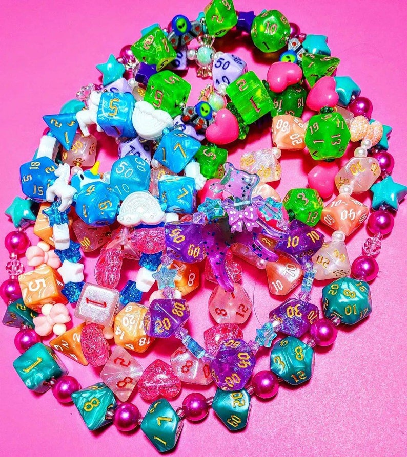 Op de afbeelding: Een kleurrijke ketting gemaakt van verschillende dobbelstenen, kralen en bedels. De dobbelstenen zijn in verschillende kleuren, waaronder roze, blauw, groen en paars. De kralen zijn in verschillende kleuren, waaronder roze, wit en zwart. De bedels omvatten een vlinder, een hart en een ster.