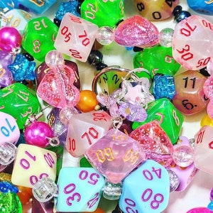 Op de afbeelding: Een kleurrijke kralenketting met verschillende dobbelsteenbedels. De dobbelstenen zijn gemaakt van helder plastic met glitter en hebben verschillende nummers op elke zijde. De kralen zijn een mix van kleuren, waaronder roze, blauw, groen en geel.