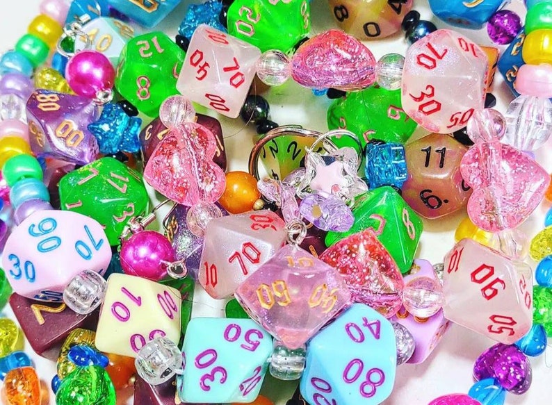 Op de afbeelding: Een kleurrijke kralenketting met verschillende dobbelsteenbedels. De dobbelstenen zijn gemaakt van helder plastic met glitter en hebben verschillende nummers op elke zijde. De kralen zijn een mix van kleuren, waaronder roze, blauw, groen en geel.