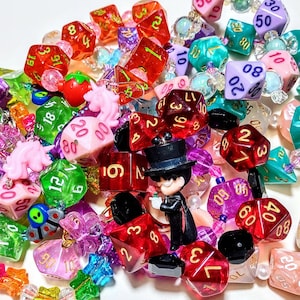 Op de afbeelding: Een verzameling kleurrijke dobbelstenen met nummers op elke zijde, sommige met glitter, sommige met een heldere plastic afwerking en sommige met een matte afwerking. De dobbelstenen zijn aan elkaar geregen met kralen en bedels, waaronder een klein beeldje van een persoon met een hoge hoed.