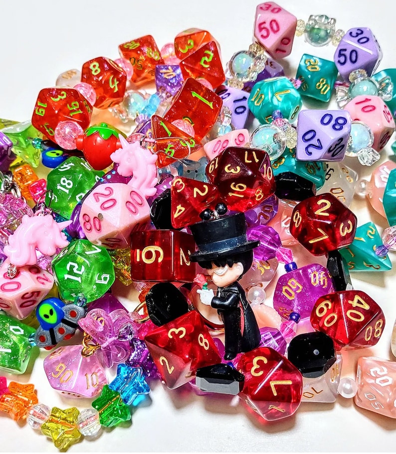 Op de afbeelding: Een verzameling kleurrijke dobbelstenen met nummers op elke zijde, sommige met glitter, sommige met een heldere plastic afwerking en sommige met een matte afwerking. De dobbelstenen zijn aan elkaar geregen met kralen en bedels, waaronder een klein beeldje van een persoon met een hoge hoed.