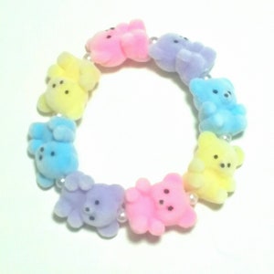 K&ouml;nnte beinhalten: Ein pastellfarbenes Armband mit einer Reihe kleiner Teddyb&auml;r-Anh&auml;nger. Die B&auml;ren sind in den Farben Rosa, Gelb, Blau und Lila.
