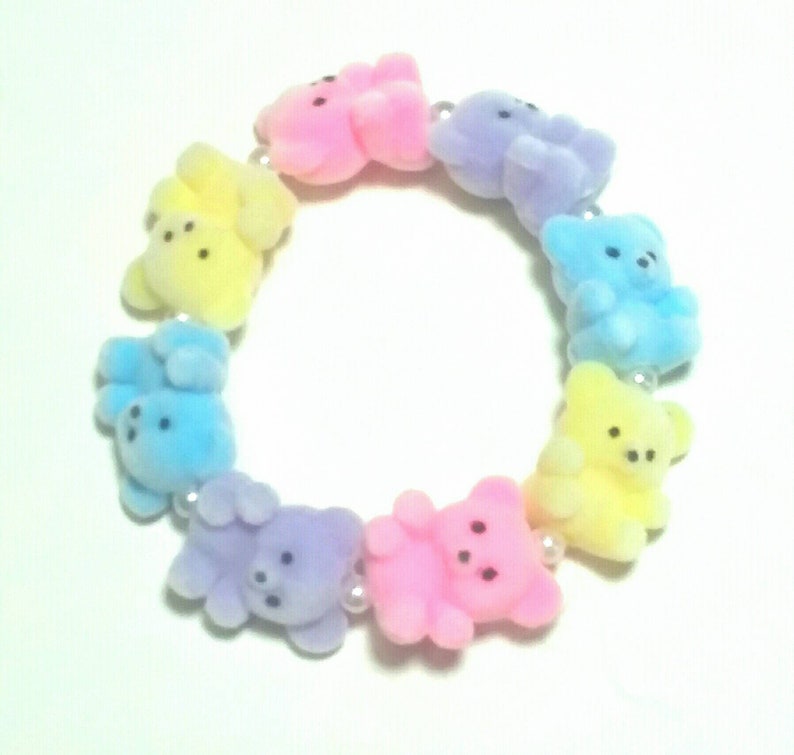 K&ouml;nnte beinhalten: Ein pastellfarbenes Armband mit einer Reihe kleiner Teddyb&auml;r-Anh&auml;nger. Die B&auml;ren sind in den Farben Rosa, Gelb, Blau und Lila.
