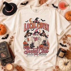May include: A cream-colored short-sleeve t-shirt featuring a graphic print of three ghosts wearing witch hats and sunglasses. The text reads "LOCAL COVEN SOCIAL CLUB" with "SPOOKY FRIENDS + GOOD DRINKS + MAGIC ALWAYS" below. The design includes pumpkins, books, and a disco ball.