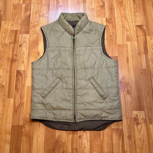 May include: A sleeveless quilted vest in a muted olive green color featuring a high collar, a full-length central zipper, and two slanted front pockets. The garment has a dark brown interior lining visible at the collar and the bottom hem edge.