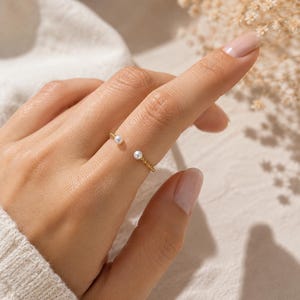 Puede incluir: Un anillo abierto de oro con un diseño de banda trenzada y texturizada. Los dos extremos del anillo terminan cada uno con una pequeña perla blanca redonda, creando una apariencia delicada y equilibrada en el dedo.