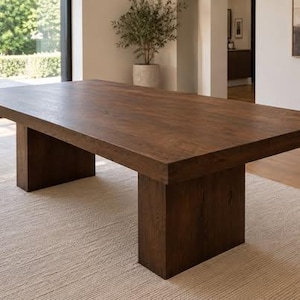 Peut inclure: Une grande table à manger rectangulaire fabriquée en bois brun foncé avec un grain visible. La table présente une surface de plateau épaisse et plate, soutenue par deux pieds solides et robustes en forme de bloc à chaque extrémité.