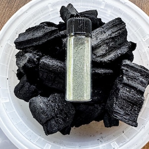 May include: A clear plastic container filled with irregular, porous black charcoal chunks. Centered on top of the charcoal is a small, upright, cylindrical glass vial with a black ribbed screw cap, containing a fine, light-colored powdery substance.